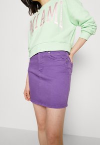 Felpa verde chiaro a maniche lunghe con "MIAMI" in lettere rosa abbinata a una gonna di jeans viola aderente. La gonna presenta tasche e chiusura con bottone.