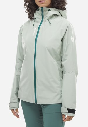 Femme portant une veste imperméable gris clair avec capuche et fermeture éclair turquoise, assortie à un pantalon turquoise, vue du cou aux hanches.