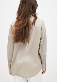 Helles beige Langarmhemd mit Knöpfen, das eine glatte Textur hat, abgerundetem Saum und Manschetten mit braunen Knöpfen. Der Stoff scheint leicht zu sein.
