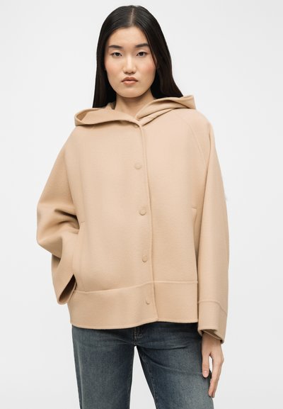 Γυναίκα με μακριά μαύρα μαλλιά που φοράει μπεζ σακάκι με κουκούλα και κουμπιά και σκούρο μπλε τζιν, στέκεται με το ένα χέρι στην τσέπη.