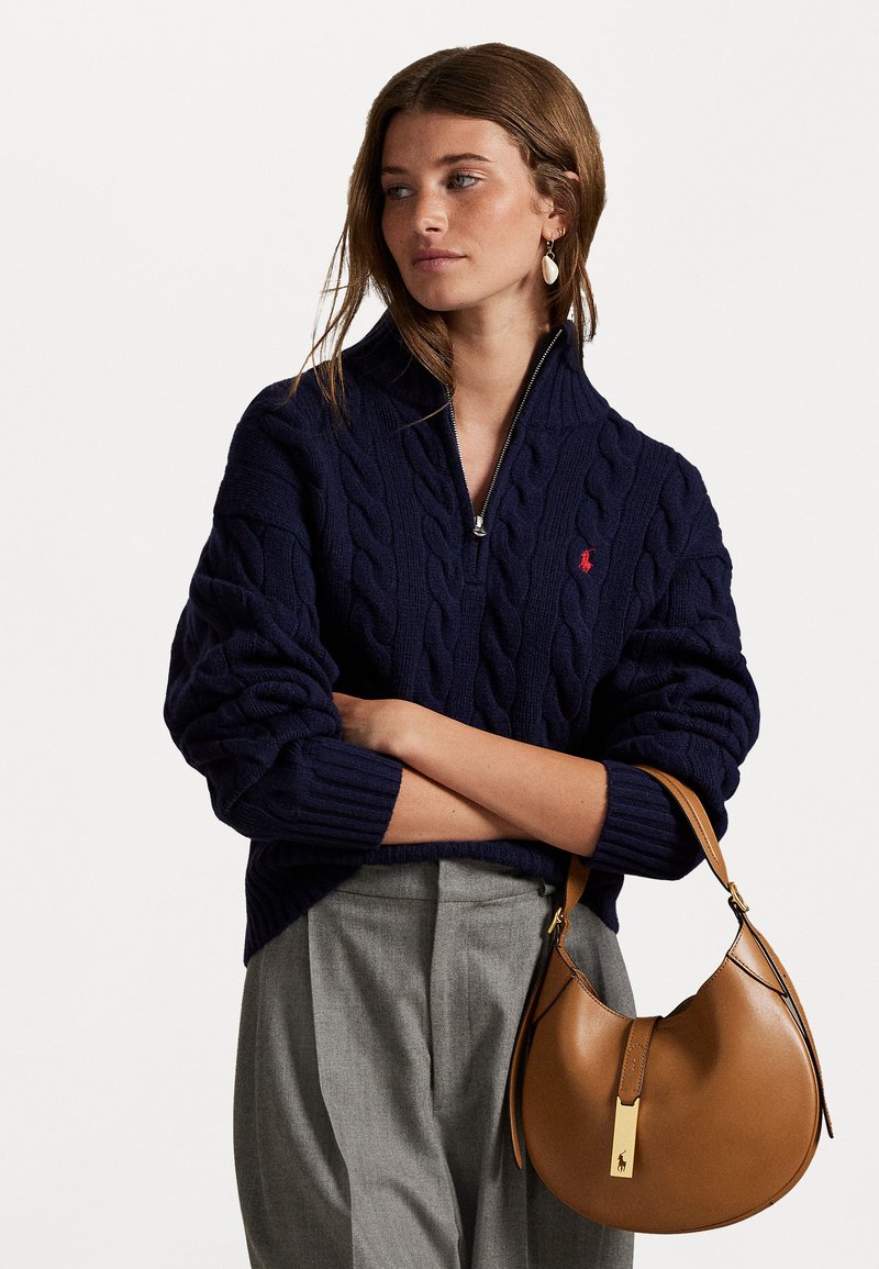 Navy cable-knit pullover with a zip neck, featuring a small red logo. Accompanied by a tan leather hobo bag with a gold accent.