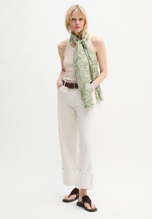 Kobieta w beżowej bluzce bez rękawów, białych spodniach o szerokich nogawkach z mankietem, brązowym pasku, zielonym szaliku w wzory związanym na szyi i ciemnobrązowych sandałach stojąca.