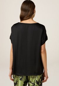 Sort satin top med korte ærmer, der har en rundet halsudskæring og glat tekstur, vist bagfra, parret med grønne mønstrede bukser.