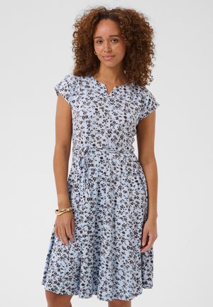 Femme aux cheveux bouclés porte une robe fleurie bleu clair à manches courtes avec une ceinture à nouer, debout devant un fond blanc uni.