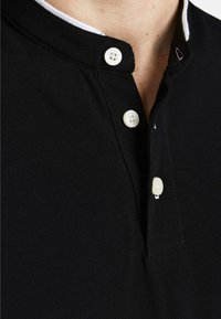 Zwarte polo met een staande kraag, twee witte knopen en een gestructureerde stof. Heeft een contrasterende witte binnenkraag.