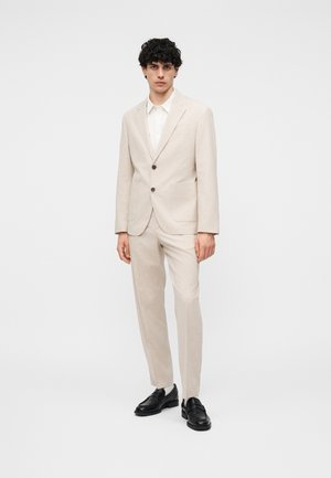 Ung mand står iført en beige jakkesæt med to knapper, hvid skjorte og sorte loafers, foran en ensfarvet hvid baggrund.