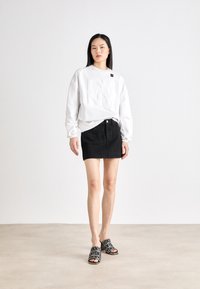 Fehér pulóver, amelyen domború "DKNY" logó található, fekete farmer mini szoknyával és mintás papucsokkal, amelyek a "DKNY" márkát viselik.