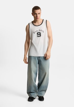 Młody mężczyzna stoi, ubrany w białą koszulkę bez rękawów z napisem „Prosto Infinity 9”, jasnoniebieskie szerokie jeansy i czarne tenisówki.