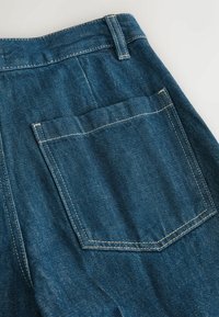 Poche arrière d'un jean en denim bleu avec coutures blanches visibles et passants de ceinture sur un fond clair.
