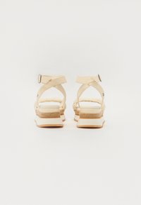 Beige sandaler med kryssremdesign, flettede detaljer rundt hælen og en platåsåle med kontrasterende striper.