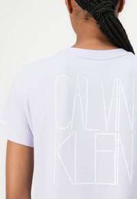 Lavanda pamučna majica s okruglim izrezom, kratkim rukavima i velikim bijelim natpisom "CALVIN KLEIN" otisnutim na leđima.