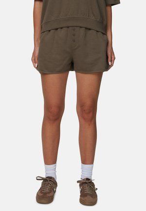 Osoba nosząca brązowe szorty na guziki, białe skarpetki do kostek oraz brązowe sznurowane trampki, stojąca na jednolitym tle.