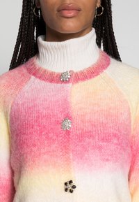 Maglione lavorato a maglia con colletto alto a costine, una gradazione di colori rosa e giallo, e tre spille decorative sul davanti.