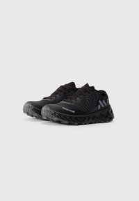 Une paire de chaussures de sport noires avec des semelles texturées, des motifs de lignes blanches ondulées et des logos de marque sur les côtés et les bouts, sur un fond gris uni.