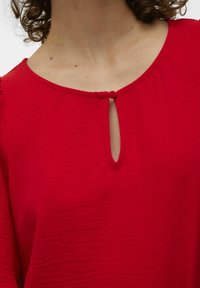 Rød top med rund halsudskæring og en lille nøglehul detalje, dropped skuldersøm og et struktureret stof med en let glans.