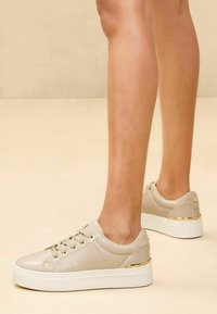 Beige bőr sneakers fehér platform talppal, arany kiegészítőkkel, lekerekített orral és klasszikus fűzős kialakítással.
