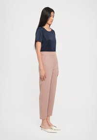 Donna in piedi di profilo che indossa una blusa blu navy a maniche corte, pantaloni rosa a vita alta e décolleté a punta color crema su sfondo bianco.