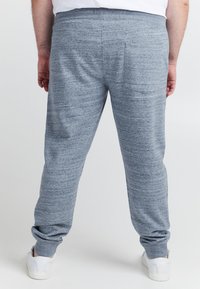 Grijze melange trainingsbroek met elastische tailleband, taps toelopende pijpen en geribde boorden. Draagt wit sneakers, gladde textuur, minimale details.