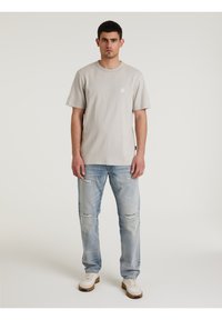 T-shirt azzurro chiaro a maniche corte con un patch quadrato sul petto, abbinato a jeans chiari strappati e sneakers bianche. Texture piatta.