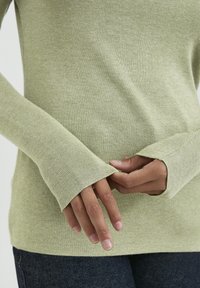 Haut à manches longues côtelé vert clair avec des poignets allongés. Gros plan sur des mains tenant le tissu, mettant en valeur les détails de texture et d'ajustement.