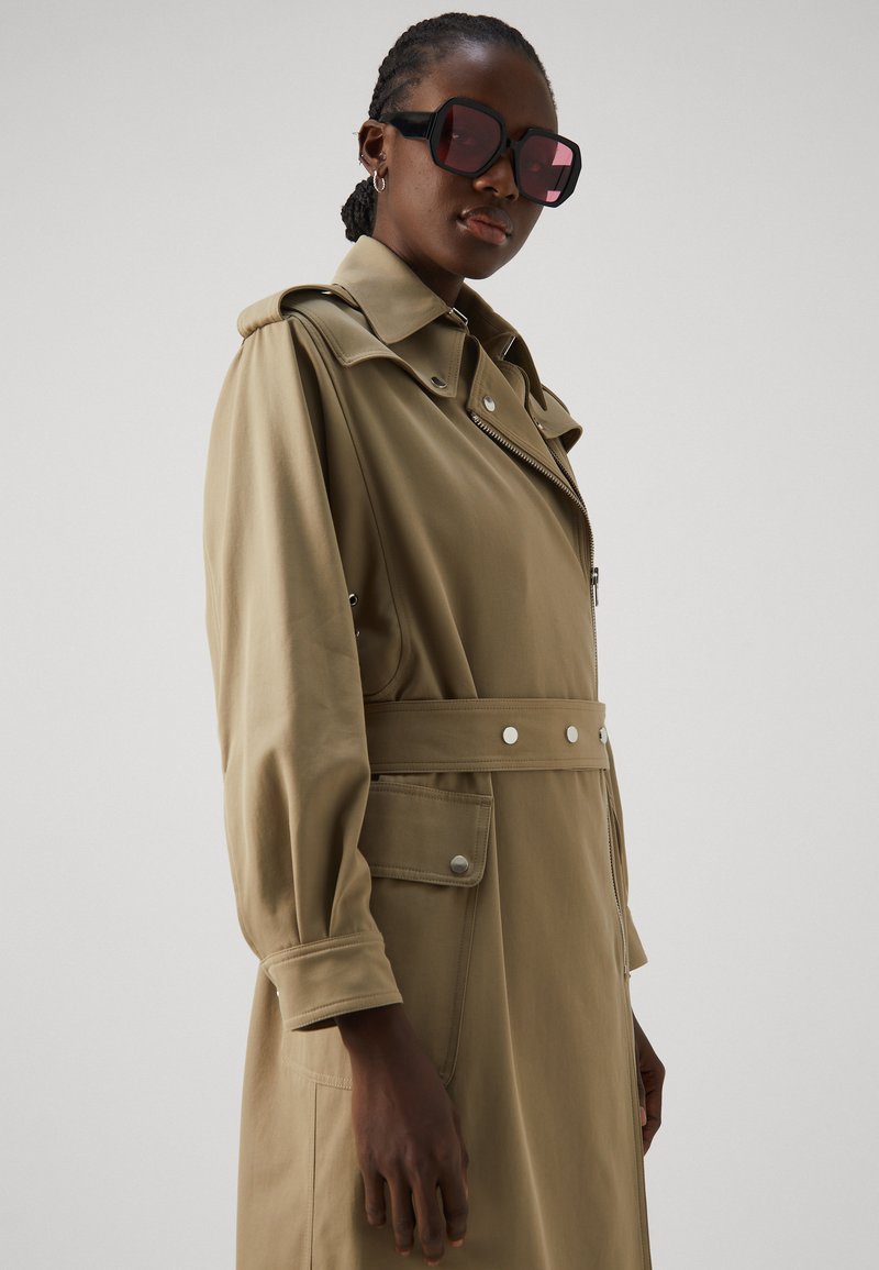 Khakifarbener Trenchcoat mit tailliertem Gürtel, oversized Kragen und Druckknöpfen. Er bietet lange Ärmel und einen Reißverschluss vorne. Dunkle Sonnenbrille mit rosa Gläsern.