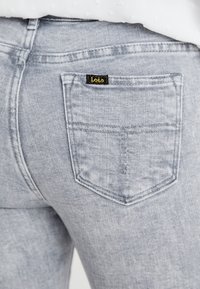I jeans grigi chiari presentano una particolare tasca posteriore cucita con un'etichetta nera e gialla che riporta la scritta "Lois". Il tessuto mostra una texture sbiadita.