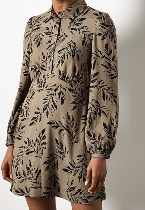 Beżowa sukienka z długim rękawem w czarne liściaste wzory, zapięta na guziki przy kołnierzyku, delikatnie marszczona w talii, z elastycznymi mankietami, noszona przez stojącą osobę.