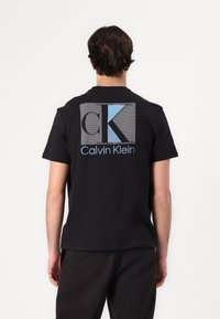 Black cotton t-shirt with short sleeves, featuring a large CK logo in blue and gray on the back. Simple, relaxed fit design.