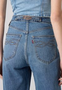 Jean en denim bleu à taille haute, avec deux poches arrière et des coutures décoratives en motif infini. Étiquette Levi's standard visible.