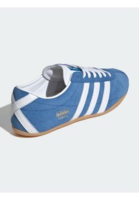Scarpa da ginnastica in camoscio blu con strisce bianche e design con lacci. Presenta un logo dorato con la scritta "adidas TOKYO" sul lato e una suola in gomma color caramello.