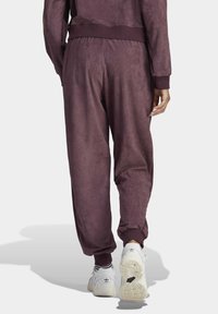 Lila, texturerade joggers med elastisk midja och mudd. Bärs med vita sneakers som har en svart logotyp. Bakvinkel, enfärgad bakgrund.