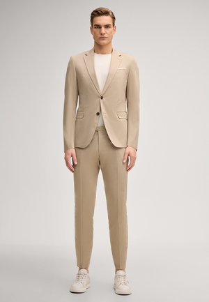 Mężczyzna stojący w beżowym, dopasowanym garniturze z białą koszulą i beżowymi sneakersami na jednolitym jasnym tle.