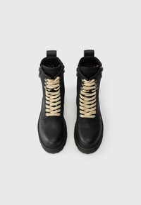 Črne usnjene gležnarje z bež vezalkami, okroglo konico, teksturirano površino, stranski zadrgo in zanko na peti. Ravna gumijasta podplat.