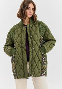 Žena oblečená v nadměrné zelené prošívané bundě s vzorovanými bočními panely a texturovanými pletenými rukávy, stojící s rukama v kapsách.