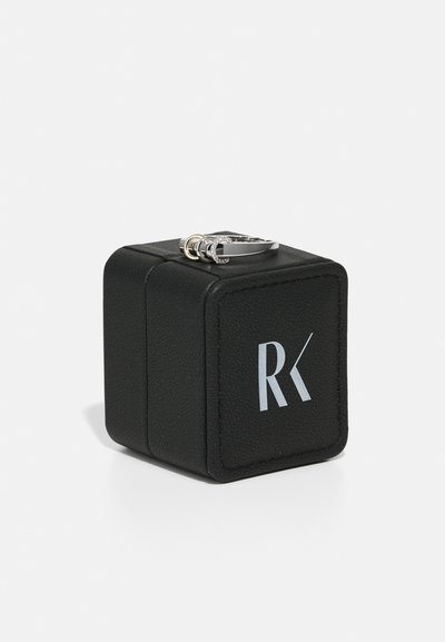 Melnas ādas gredzenus kastīte ar tekstūru, priekšpusē ar sudraba logo "RK". Izliek sudraba gredzenu ar trim savienotām joslām.