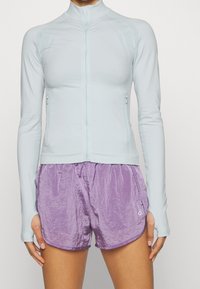 Světle modrá dlouhá sportovní bunda s zipovým uzávěrem a texturovanými panely, spárovaná s fialovými kraťasy s pomačkaným vzhledem.
