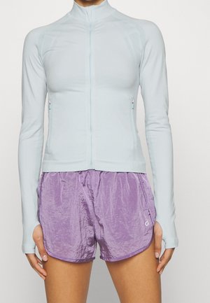 Lichtblauwe sportjack met lange mouwen, voorzien van een ritssluiting en tekstuuroverlays, gecombineerd met paarse shorts met een gewrinkeld aanzien.