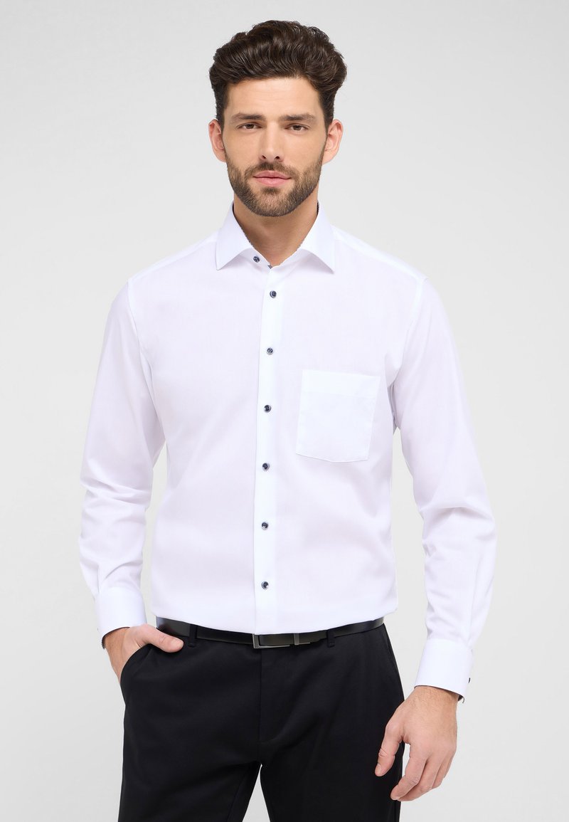 Camicia bianca a maniche lunghe con colletto button-down, bottoni neri a contrasto e taschino sul petto sinistro, abbinata a pantaloni neri.