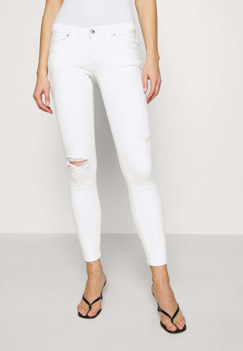 Femme portant un jean slim blanc déchiré et des sandales à talons noires, debout devant un fond clair uni.