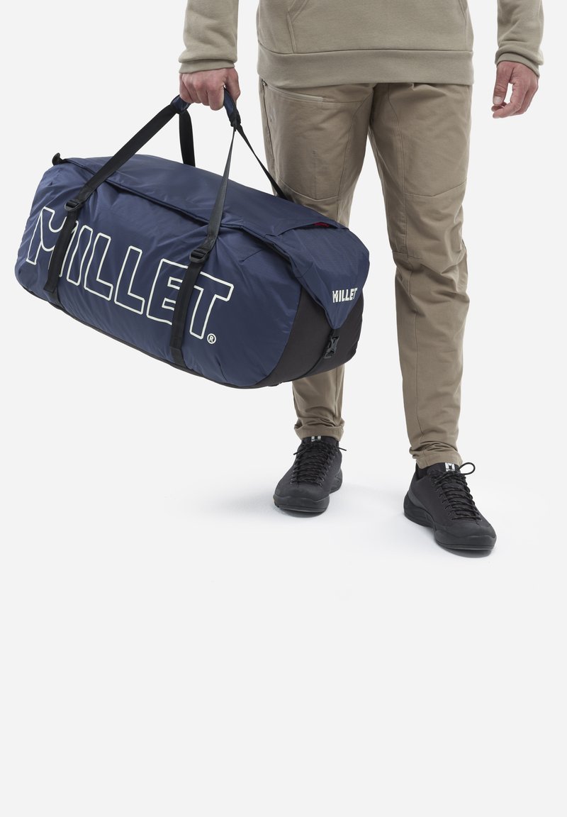 Sac de sport bleu avec le logo blanc "Millet", sangles en tissu et une fermeture supérieure sécurisée, tenu par une personne en pantalon beige et chaussures noires.
