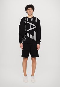 Felpa nera con grande logo grigio e strisce, abbinata a pantaloni corti neri. La modella indossa una piccola borsa a tracolla nera e sneaker bianche.