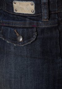 Jeans in denim scuro con tasca frontale con chiusura a bottone. Etichette visibili con dettagli di cucitura e filo a contrasto sottile.