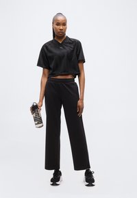 Γυναίκα με μαύρα αθλητικά ρούχα κρατώντας ένα διαφανές μπουκάλι νερού με μαύρα και λευκά αθλητικά παπούτσια, στέκεται μπροστά σε έναν απλό ανοιχτόχρωμο φόντο.