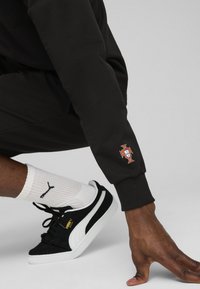 Sort sweatshirt med Portugal-flag emblem på håndleddet. Person bærer sorte og hvide PUMA sneakers samt hvide sportsstrømper med mønster.