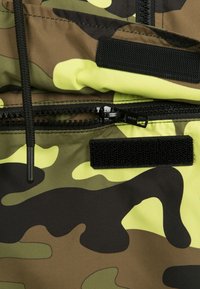 Kamouflage-mönstrad tyg i nyanser av grönt, svart och neongult med dragkedjestängning, justerbara snörningar och kardborreaccenter.
