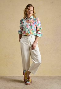 Femme portant une chemise fleurie à manches longues, un pantalon blanc taille haute à jambes larges et des mocassins multicolores, posant devant un fond beige.