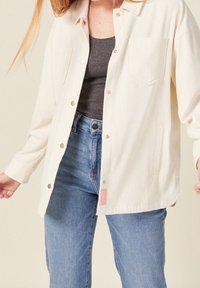 White ribbed button-up jacket with a pocket, gold buttons, and a pink brand tag; worn over a gray top and blue jeans.