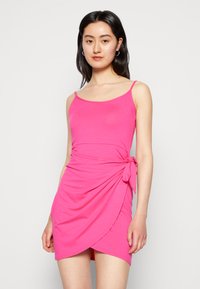 Vestido envolvente rosa feito de um tecido macio e elástico, com alças finas e um detalhe para amarrar de lado. Apresenta um design drapeado e um comprimento acima do joelho.