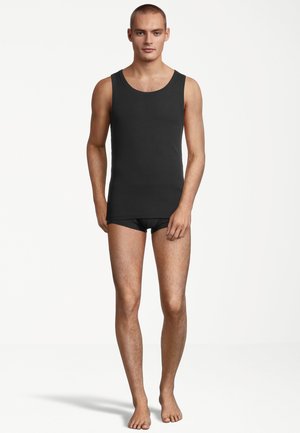 Schwarzes Tanktop aus Baumwolle mit einem runden Ausschnitt und schlanker Passform, kombiniert mit schwarzen Slips. Modell steht barfuß auf einem neutralen Hintergrund.