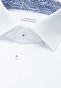 Chemise blanche avec une doublure de col à motif paisley bleu, deux boutons blancs avec une couture bleu foncé, et une finition en tissu texturé.